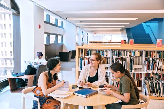 Three students sit at a round table in front of bookcases in the University Center Library