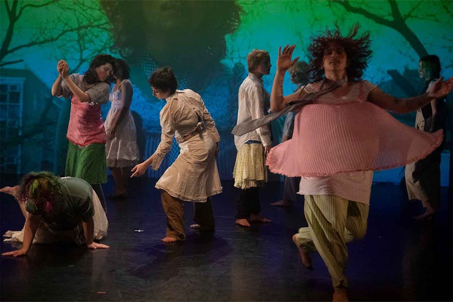 Photo of captured in different positions--some standing, crouching, twirling, clapping. A projected image behind them features a medium close-up of someone standing in front of trees and buildings.