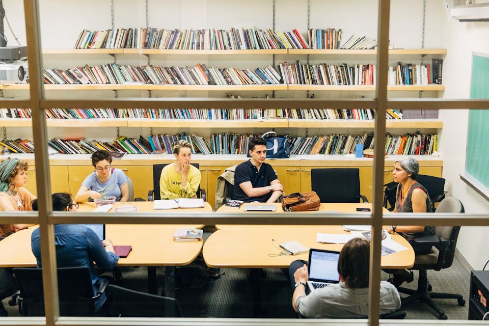 Students sit in front of a bookcase at a table in a classroom.