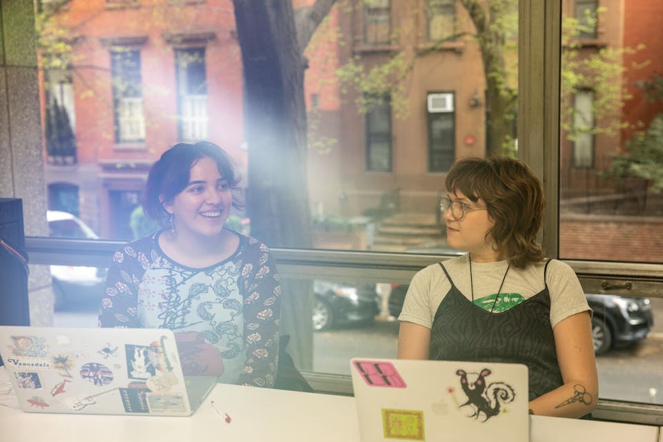 Two students sit in a classroom and talk in front of a wall of windows in the Lang building.
