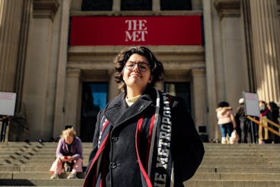 A person wearing a peacoat and glasses and standing on the steps of The Metropolitan Museum of Art is photographed from below.