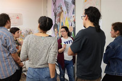 Students stand in a circle in an art studio looking at a piece of art. One student speaks.