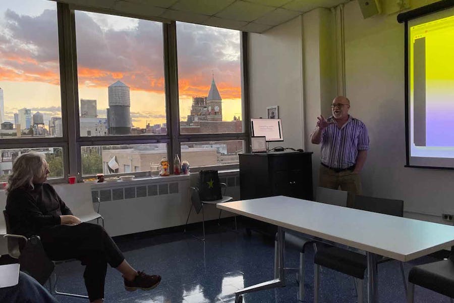 Michael Pettinger presents in a classroom with windows looking out on the sun setting in New York City.