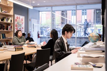 Students study with books on their tables in front of a wall of windows. A person walks by on the sidewalk outside.