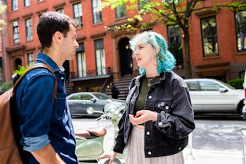 A woman with teal-colored hair talks to a friend on a tree-lined street near a red-bricked building in the city.