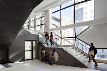 Students climb a staircase in a modern light-filled room with large glass windows overlooking mid-rise buildings outside.