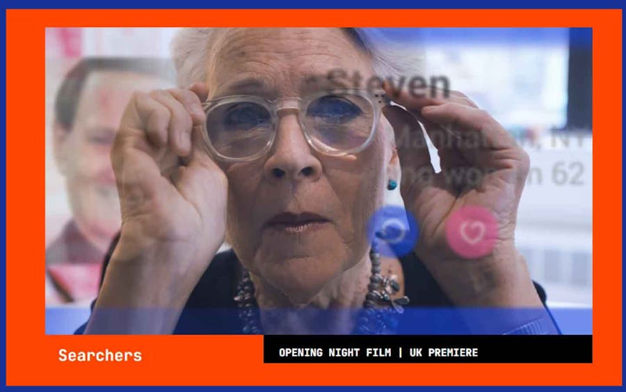 Still from the film, Searchers, showing the reflection of a woman facing the camera,  adjusting her glasses. Her image is reflected on a screen with the profile of a person named Steven.