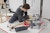 Student sitting on the floor surrounded by materials scraps and various equipment tinkers with a 3D printer.