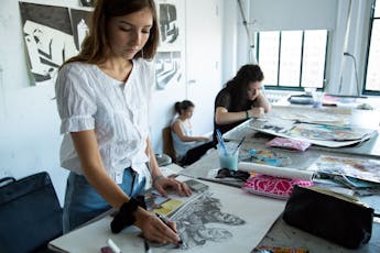 White Female Student with long brown hair and a white top and jeans draws an image of two people in pencil while standing in a classroom.  In the background are two other female students drawing.