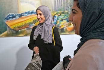 Two students smiling stand next to each other in a hallway with a painting on the wall behind them. 