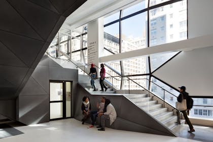 Students climb a staircase in a light-filled room with large glass windows overlooking mid-rise buildings outside.