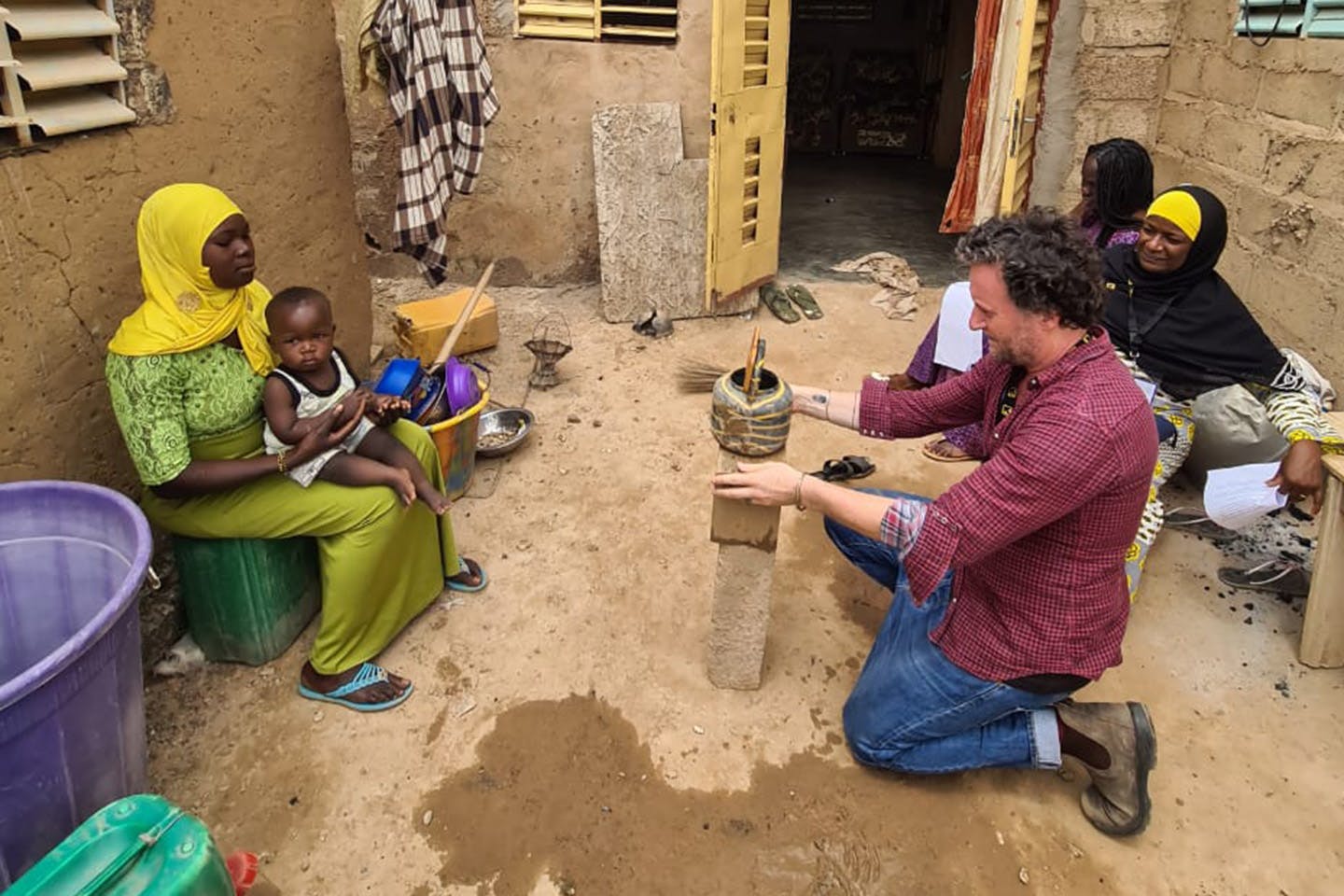 A man steadies a camera on a ceramic vessel, preparing to photograph a woman across from him, seated with a child in her arms and in colorful garb.