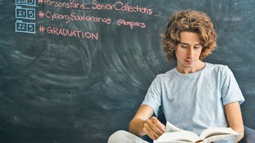 A student reads a book while sitting in front of a chalkboard with upcoming dates and hashtags. 