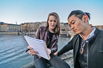 Two students read from a notebook while leaning on a concrete wall overlooking a river with buildings in the distance. 