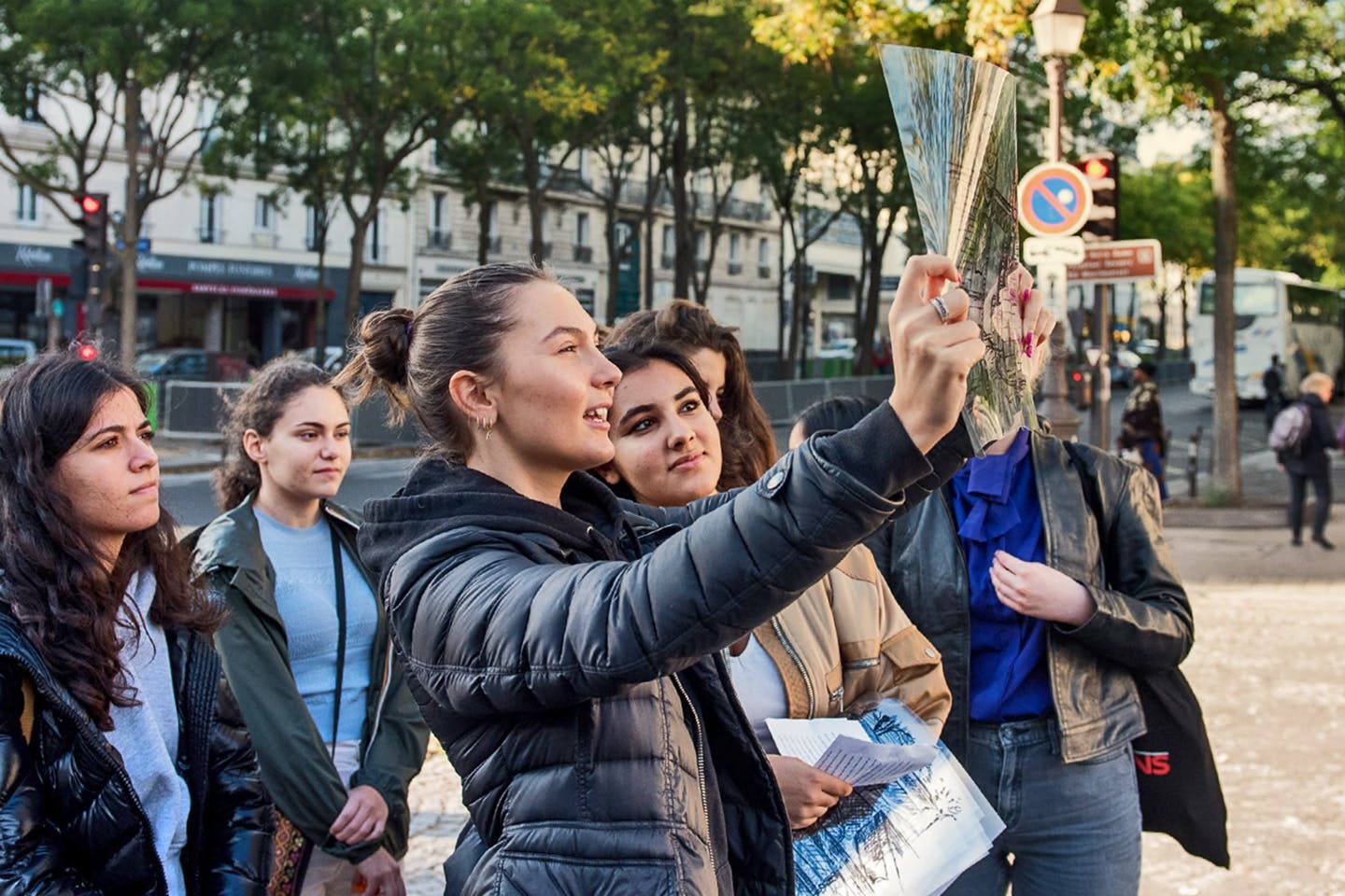 A group of students outside on the sidewalks of Paris, looking at art on a clear substrate, held up to the sun.