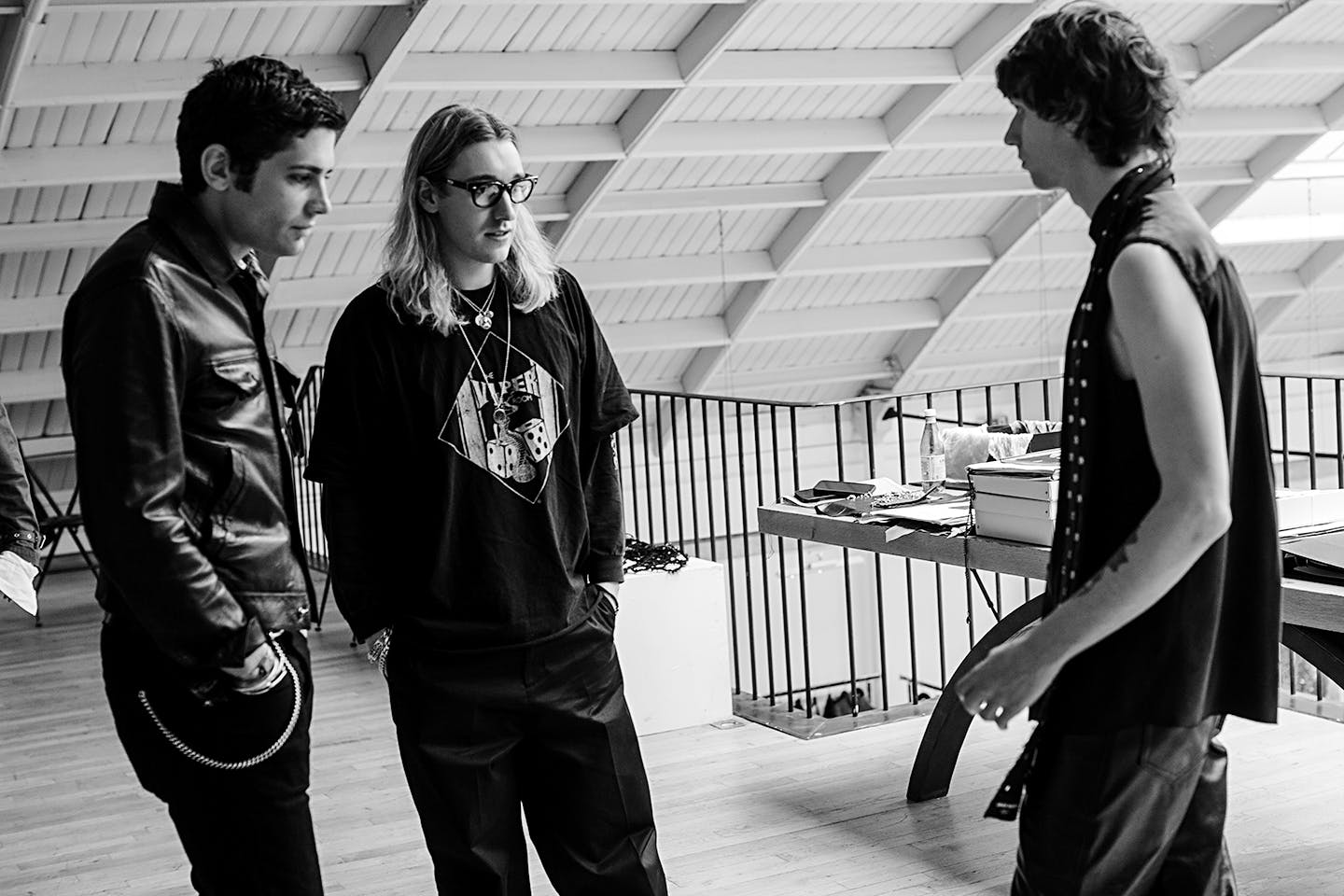 In a black and white photo, two people look at the outfit of a third in a light-filled atelier.