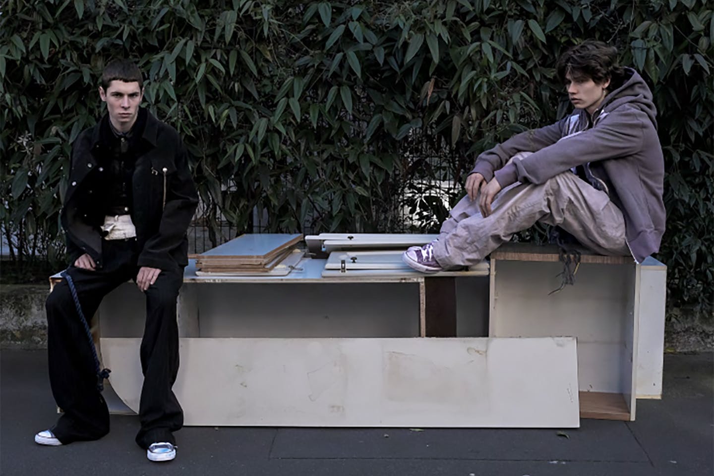 Two students sit on a broken piece of furniture outside wearing sneakers and slouchy clothing.