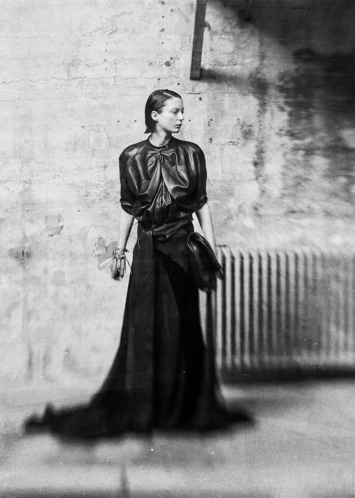 Black-and-white photograph of a model wearing a sculptural Candice Tianyu outfit with a voluminous, gathered top and long skirt, standing in a textured studio space.