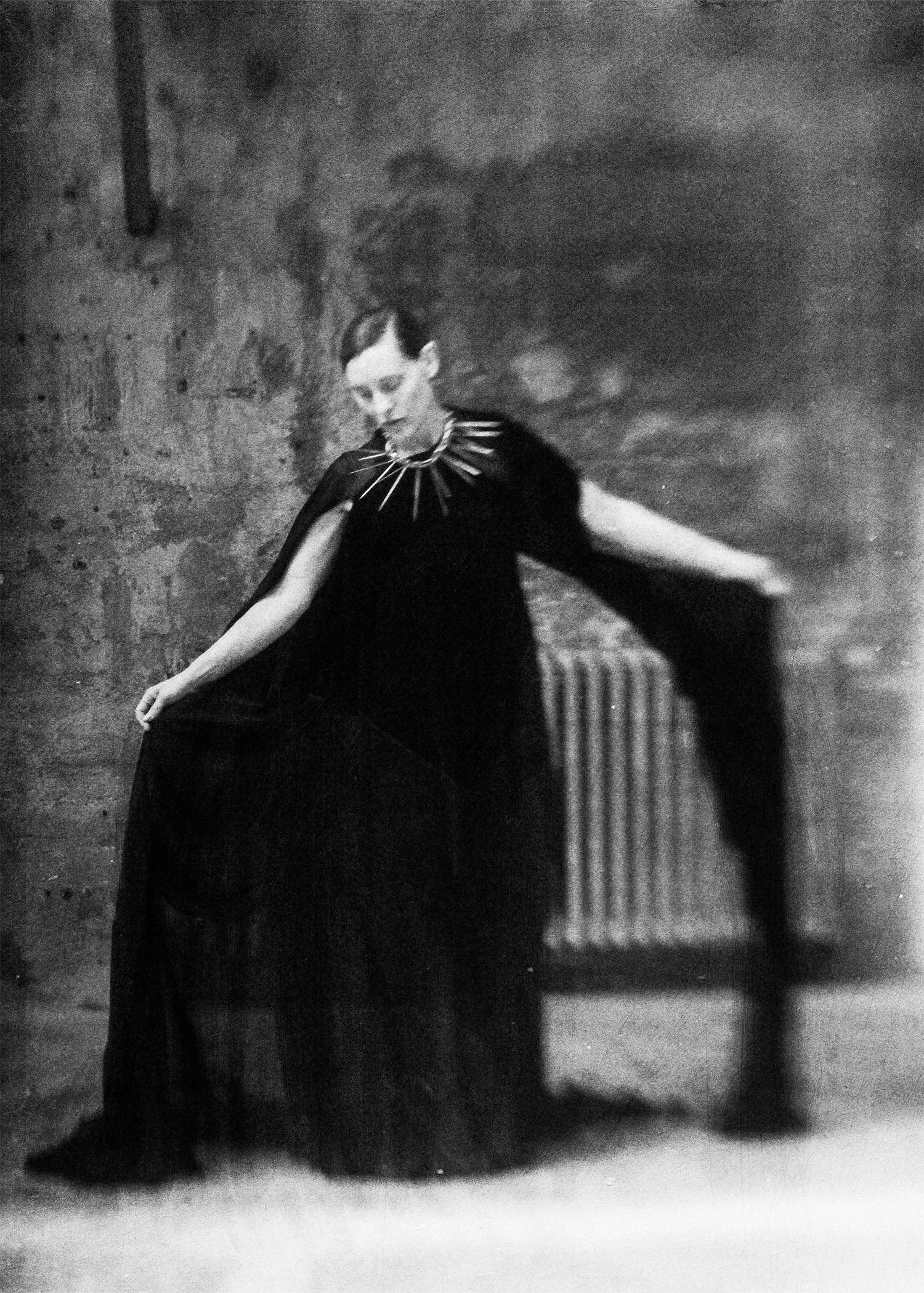 Black-and-white photograph of a model wearing a flowing black Candice Tianyu garment with a spiked, sculptural neckline, arms extended to emphasize movement.