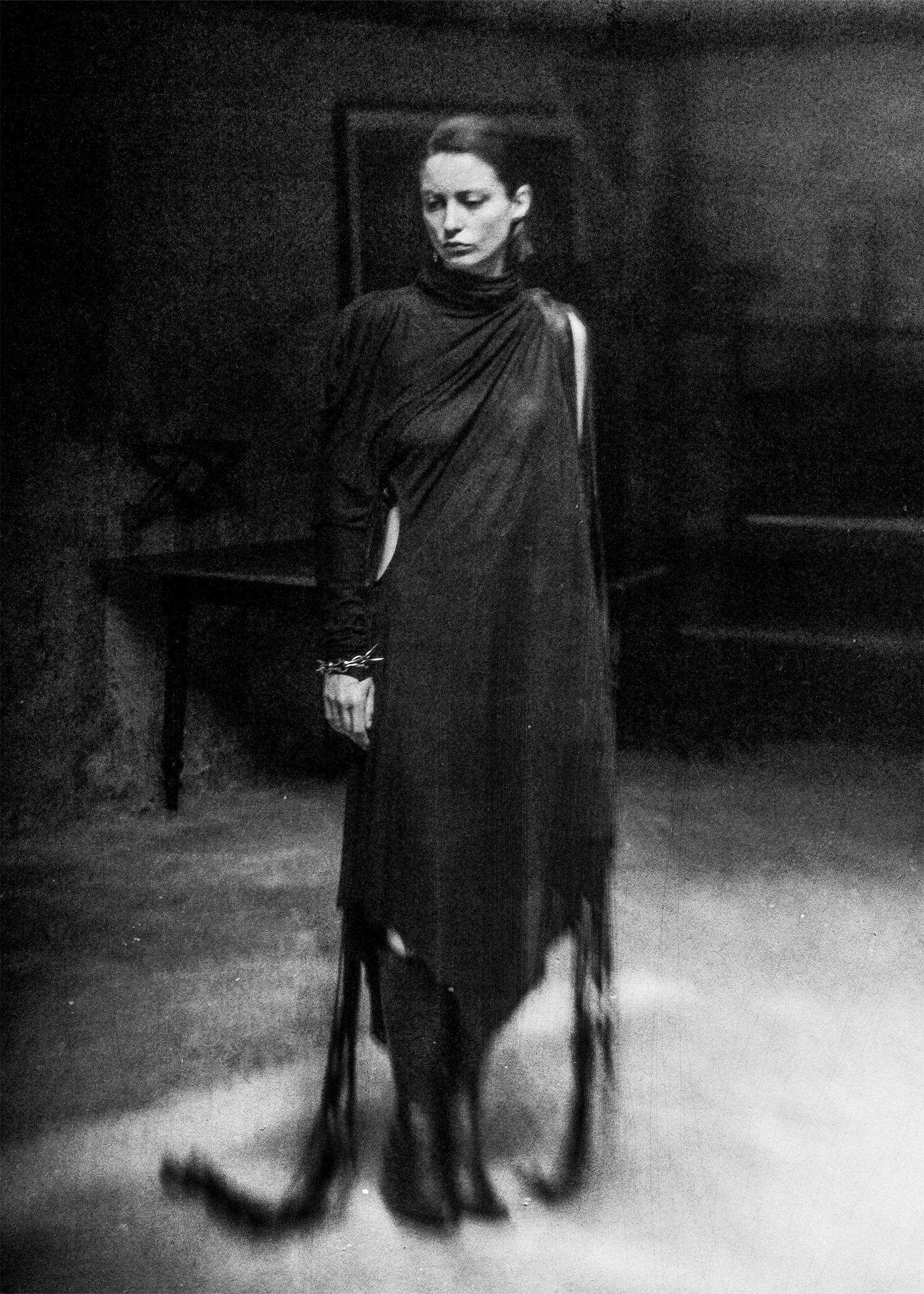 Black-and-white photograph of a model wearing a draped, asymmetrical Candice Tianyu garment with long fringe and soft cutouts, standing in a dimly lit studio.