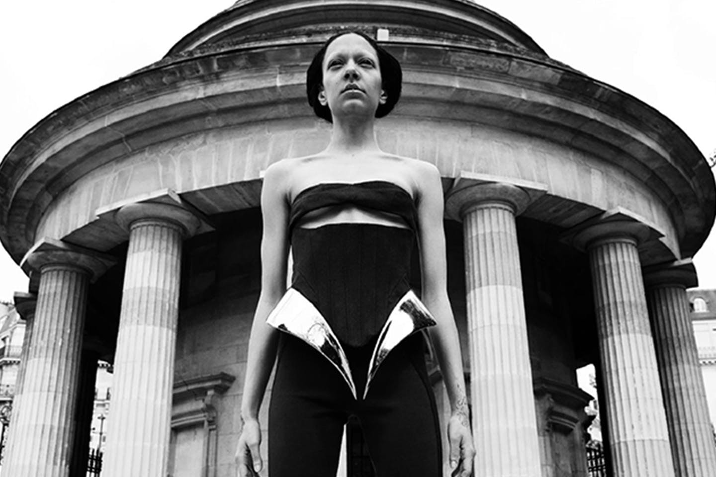 A woman stands before a neoclassical Paris building wearing a black bandeau and a pair of high-waisted pants with 3D-printed hip accents.   