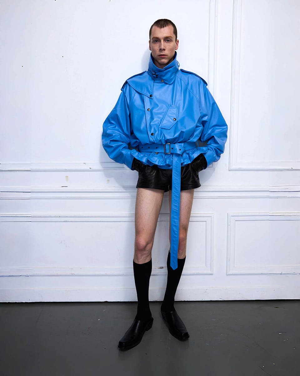 Model seated against a white wall wearing a sheer yellow textured sweater and light shorts with a black choker, designed by Xiao Xu; photographed by Boyan Li, model Paul Lemoine.