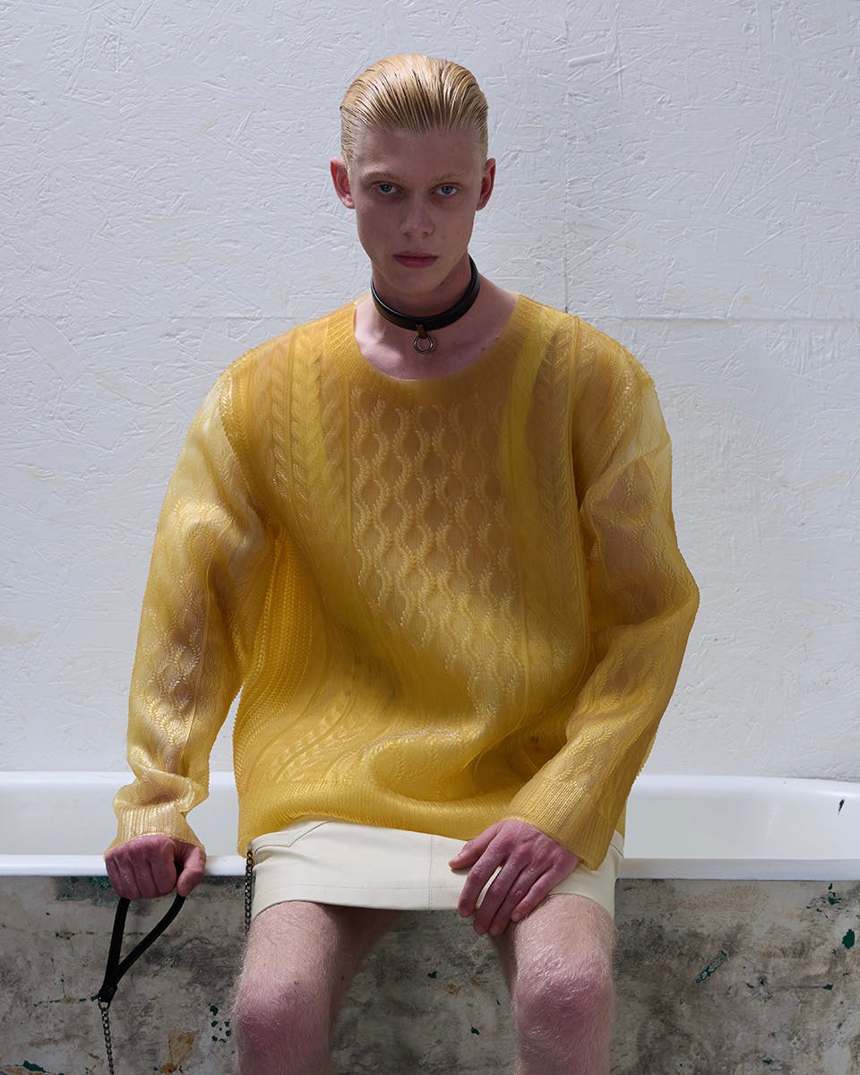 Model standing against a white wall wearing a bright blue belted jacket with black shorts and knee-high socks, designed by Xiao Xu; photographed by Boyan Li, model Matéo Picard.