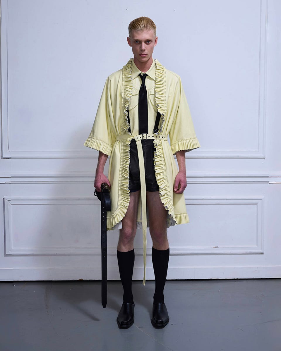 Model standing against a white wall wearing a pale yellow pleated coat with a belt, black tie, black shorts, and knee-high socks, holding a long black strap; designed by Xiao Xu, photographed by Boyan