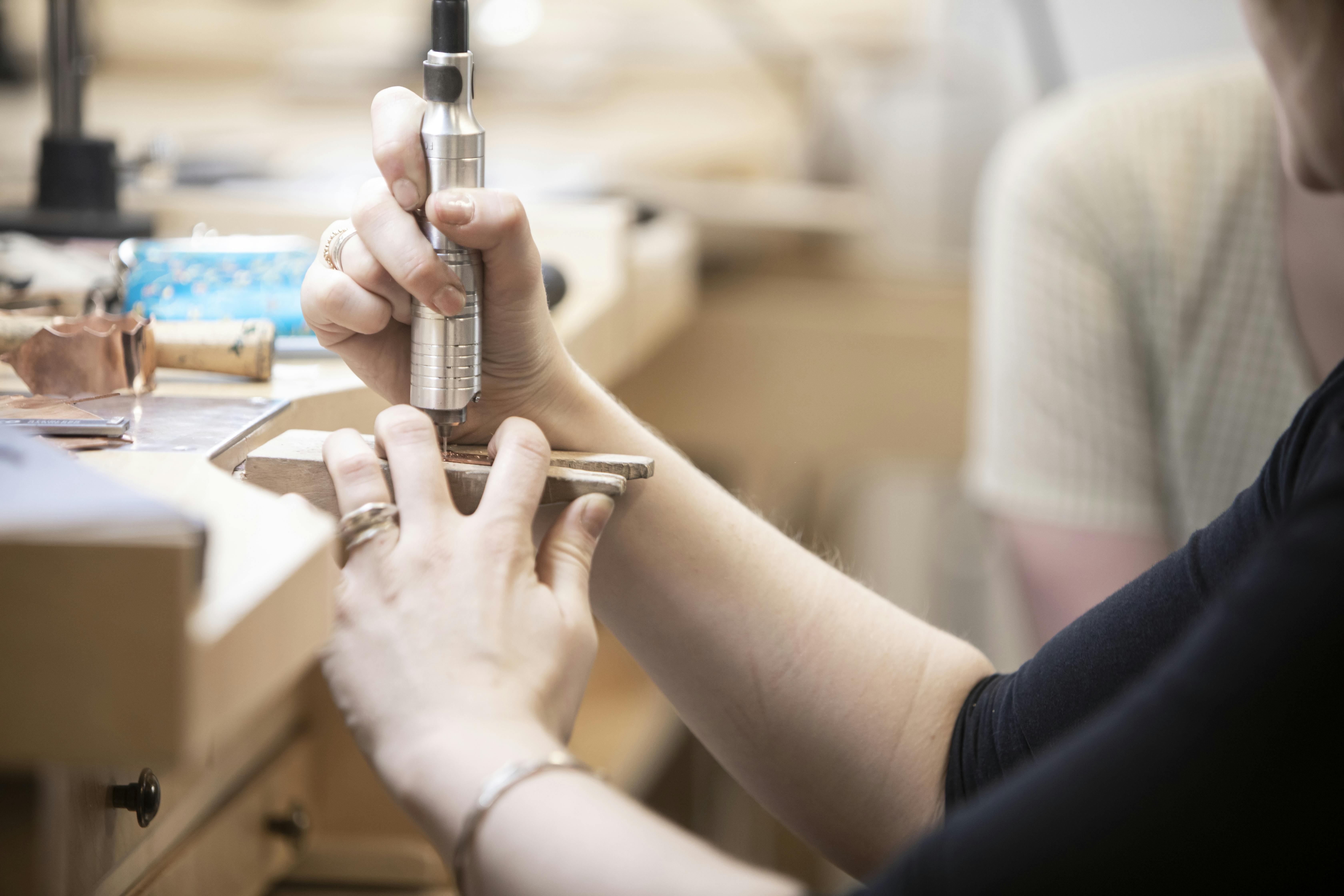 A pair of hands uses a metal tool and a wooden clamp to make jewelry.