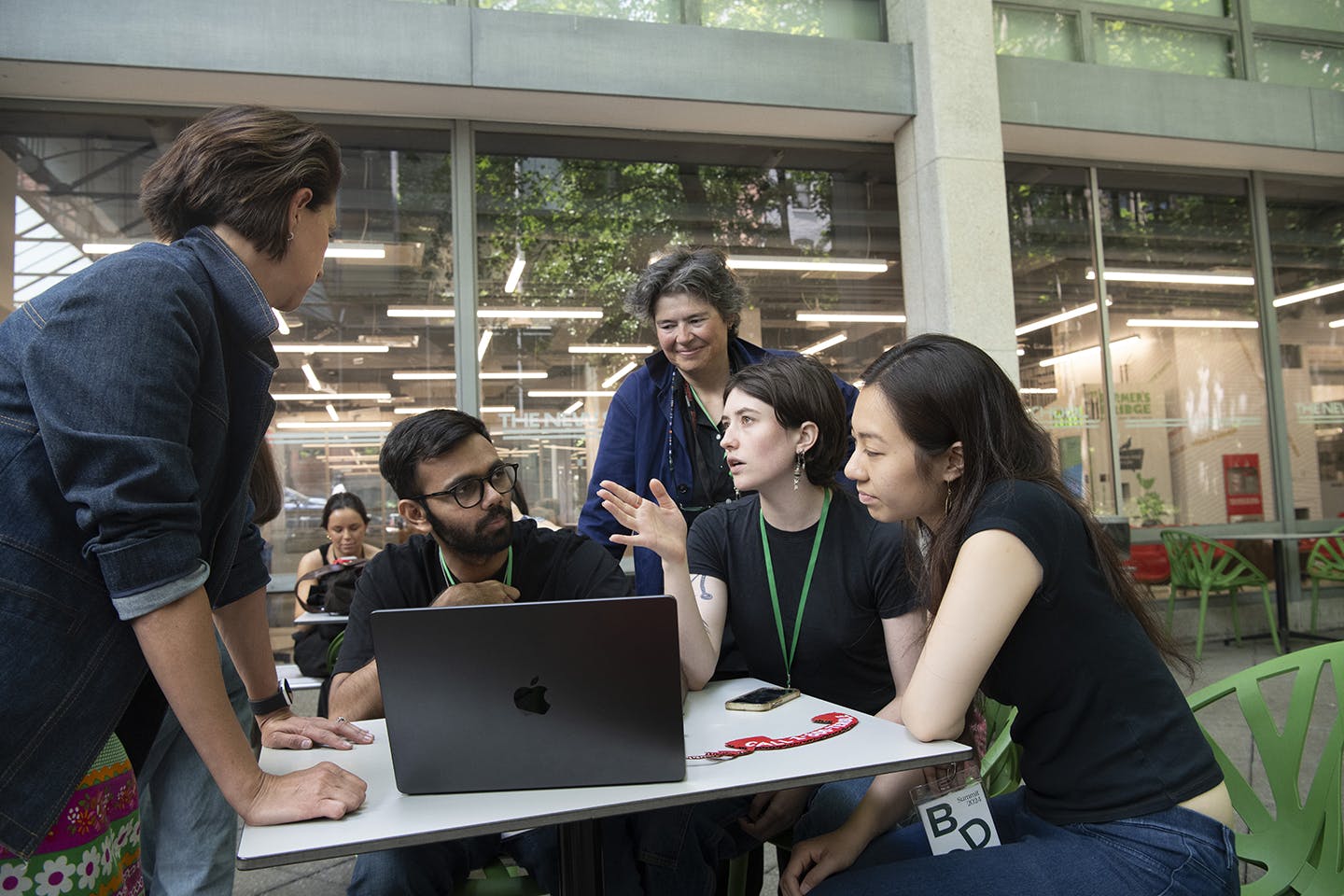 Two women stand over and observe three students, seated in an outdoor courtyard around a laptop, as they work.