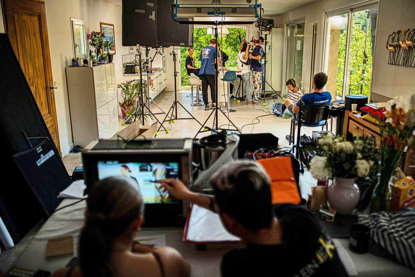 Students at a film shoot in a white room with some students in the foreground looking at a monitor, and some in the background with microphones and cameras. French doors show greenery outside.