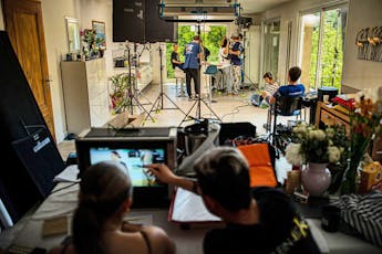 Students at a film shoot in a white room with some students in the foreground looking at a monitor, and some in the background with microphones and cameras. French doors show greenery outside.