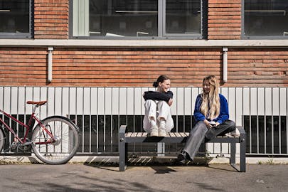 Two students talk on a bench in front of a building. There is a bicycle in the frame.