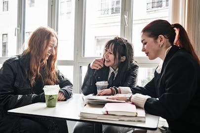 Three students study and laugh together at a table in a cafe.