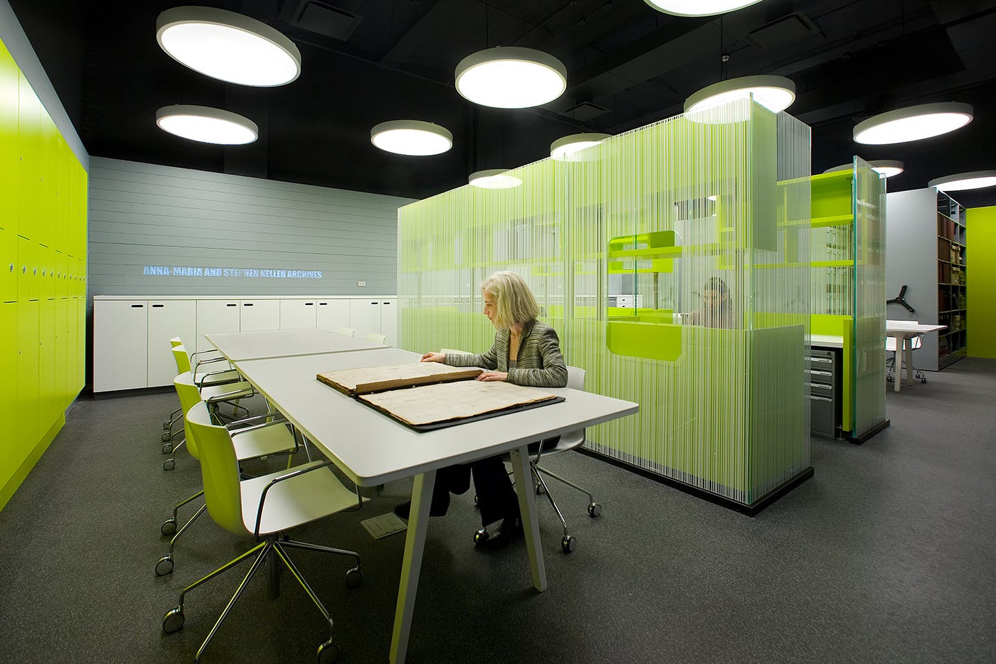 Person in a grey jacket looks at archival work on a table in a room decorated in chartreuse lockers, chairs, and desk spaces