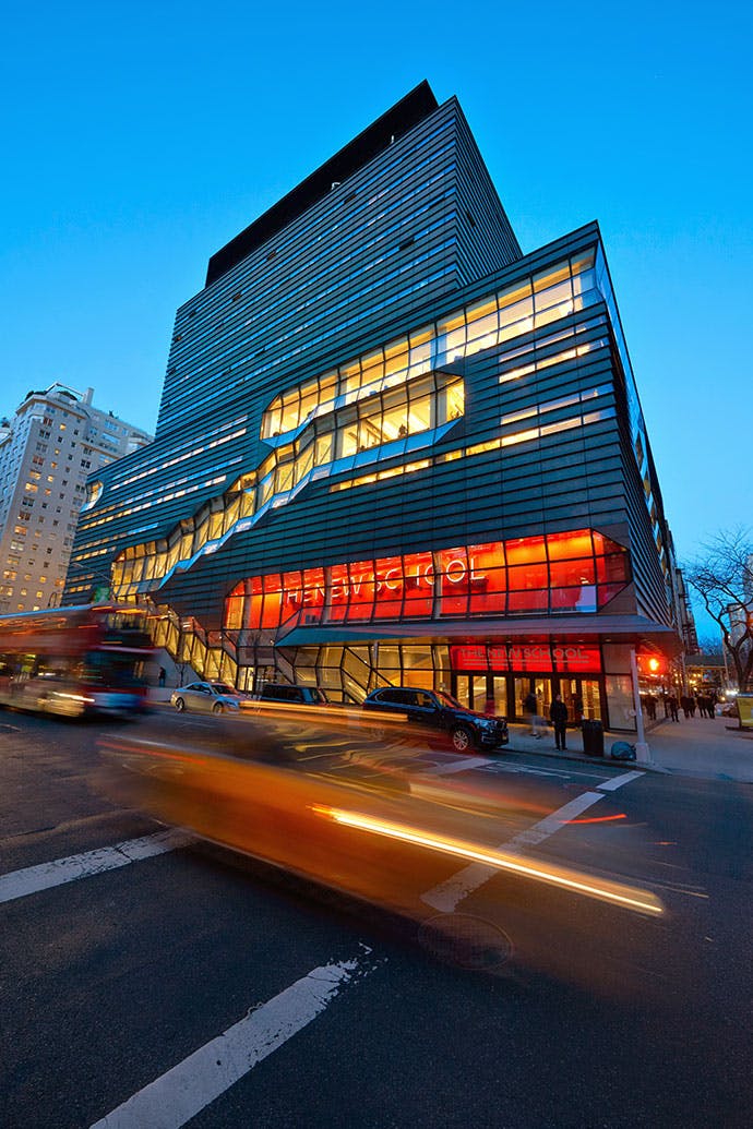 The university center in New York City's Greenwich Village, with a dramatic facade and large windows, stands on a busy street corner illuminated in the evening light. The building's orange sign with the words “The New School” can be seen through the windows.