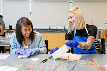 Two students in a lab smile as one shows the other their work. Both are wearing rubber gloves and working with chemicals on different fabrics.