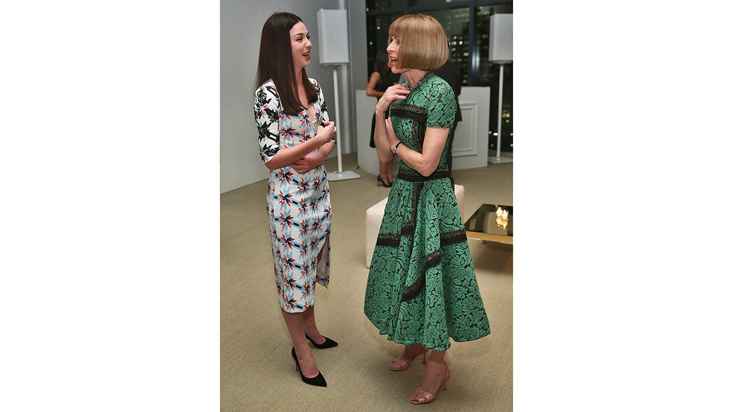 Tanya Taylor, wearing a pattered silk white dress stands speaking to Vogue magazine's Anna Wintour, wearing a green patterened dress with a window in the background showing it's night.