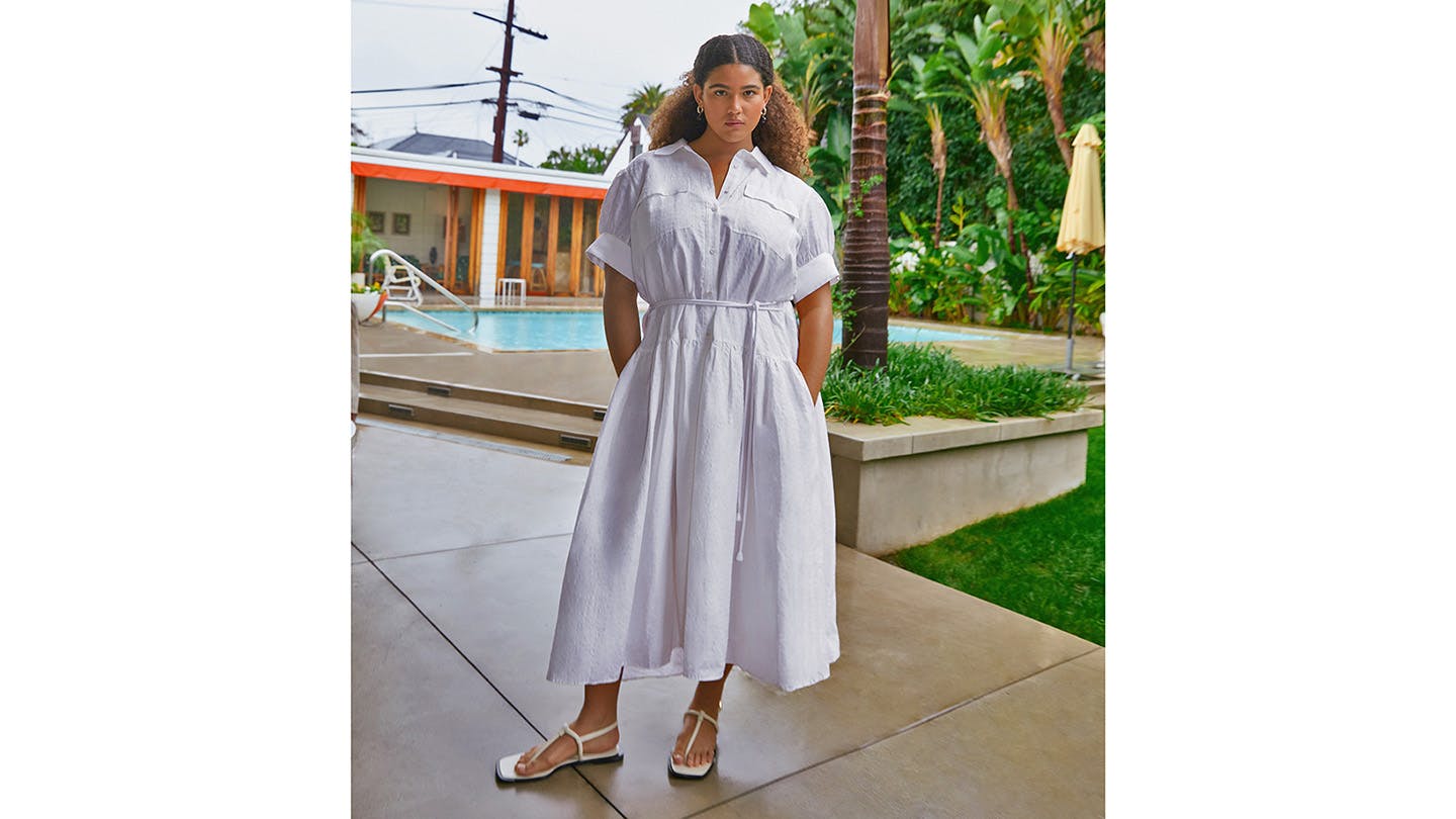 A person in a belted white dress with pockets stands in front of a swimming pool.