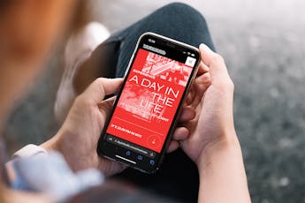 A person looks at a mobile phone in their hand which reads, in red, A Day in the Life.