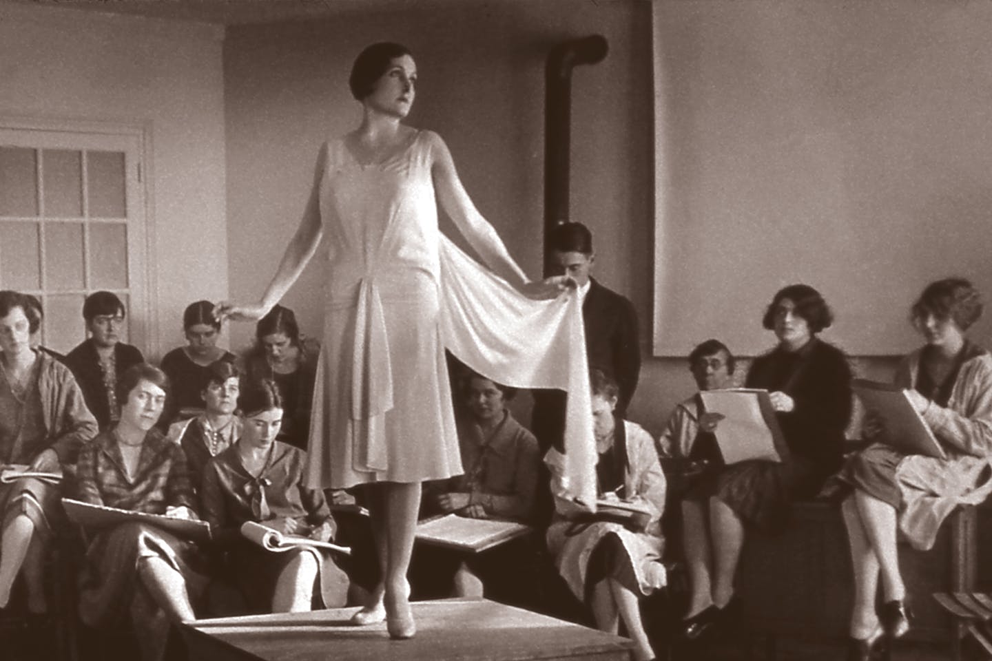 A model in a white dress poses on a platform surrounded by students drawing her. From the 1920's.