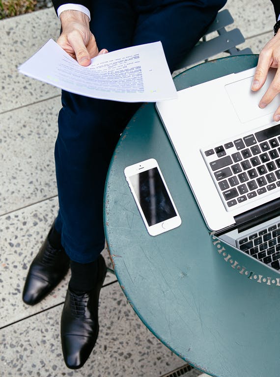 A man sits at a round table, reading a piece of paper in one hand while mousing on a laptop with his other hand