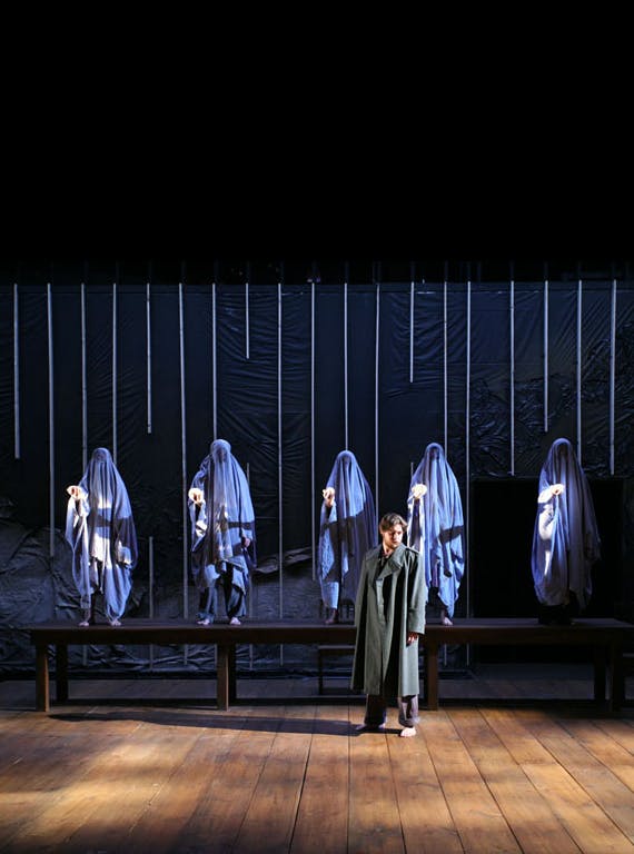Five people concealed in blue fabric stand behind an actor in a grey coat on a stage as part of the graduate program in dramatic arts in the 1990s