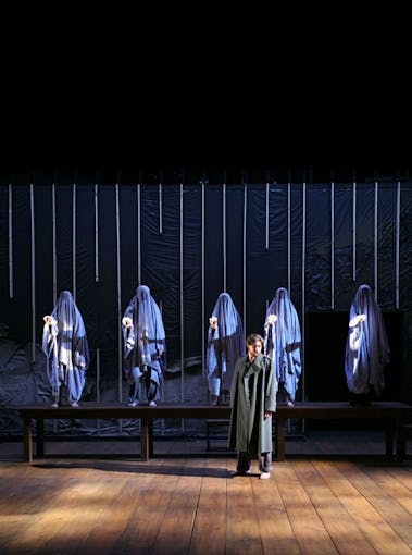 Five people concealed in blue fabric stand behind an actor in a grey coat on a stage as part of the graduate program in dramatic arts in the 1990s