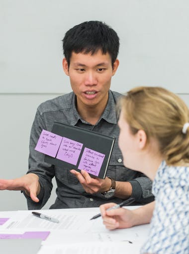 A student with sticky notes speaks to a student in a ponytail at a table