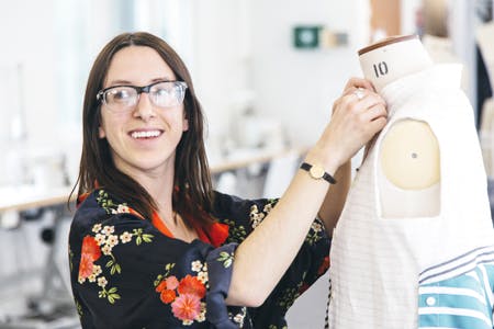 A smiling student with glasses works on a garment on a dressmaker's dummy