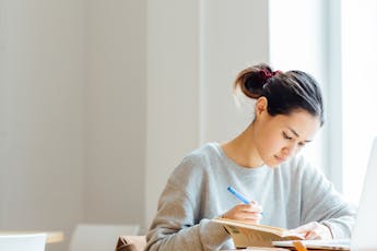 A student wearing a gray sweater makes notes on a soft-cover book in a white room.