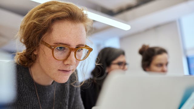 A student with red hair and brown glasses intently viewing a laptop near two out of focus students in the background