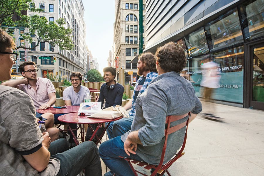A group of students sit at an outdoor cafe on a lively city street, enjoying the sunny weather and surrounded by trees and other green plants.