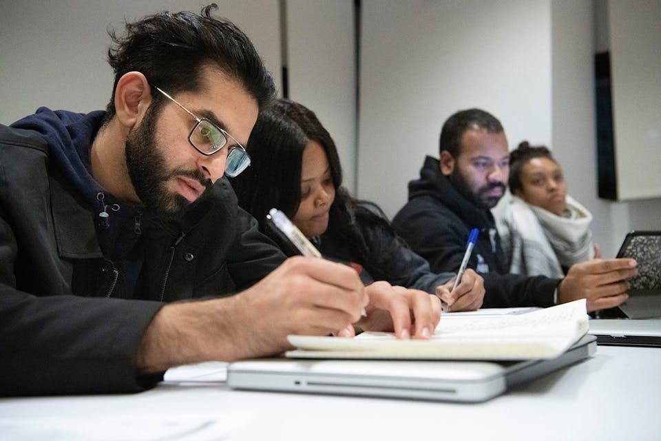 A row of four students concentrate on note taking at a desk during class.